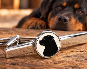 Könnte beinhalten: Eine silberfarbene Krawattenklammer aus Metall mit einer runden Vorderseite, die eine schwarze Silhouette eines Rottweiler-Hundekopfes im Profil auf cremefarbenem Hintergrund zeigt. Die Krawattenklammer hat eine polierte Oberfläche und einen Federclip.