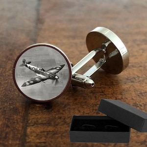 Puede incluir: Gemelos de plata con una imagen en blanco y negro de un avión vintage en una caja redonda. Los gemelos vienen en una caja de regalo negra.