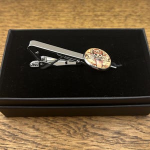 May include: A silver tie clip with a round, colourful skull and crossbones design. The tie clip is in a black velvet box.