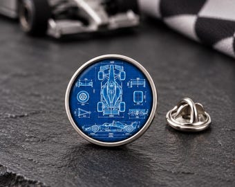 Könnte beinhalten: Eine runde Anstecknadel mit einer blau-weißen technischen Blaupausen-Illustration eines Formel-1-Rennwagens. Das Design zeigt das Auto aus der Draufsicht und von der Seite mit verschiedenen mechanischen Komponenten in weißen Linien.