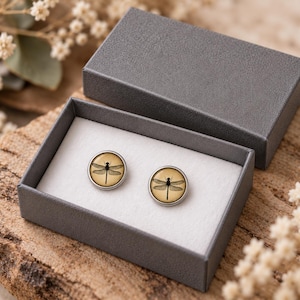 Peut inclure: Une paire de boucles d'oreilles rondes à clous présentant une illustration détaillée de libellule noire sur fond beige, présentées dans une boîte cadeau rectangulaire gris foncé avec une doublure intérieure blanche et un couvercle assorti.