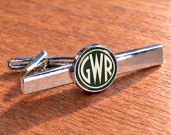 Könnte beinhalten: Eine silberfarbene Krawattenklammer aus Metall mit einem kreisförmigen Mittelstück, das einen dunkelgrünen Hintergrund und die in Cremefarbe aufgedruckten Buchstaben GWR aufweist. Die Krawattenklammer hat eine polierte Oberfläche.
