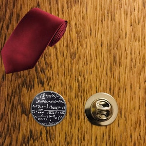 May include: A maroon tie with a silver pin featuring a mathematical equation. The pin is circular and has a black background.