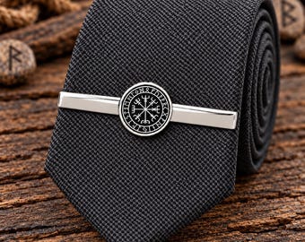 Könnte beinhalten: Eine texturierte schwarze Krawatte mit einer silberfarbenen Krawattenklammer. Die Klammer ist mit einem kreisförmigen Emblem verziert, das ein schwarz-weißes Vegvisir-Symbol zeigt, umgeben von einer Umrandung aus Runenzeichen.