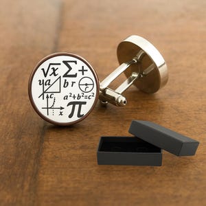 May include: Silver cufflinks with a white face featuring black mathematical symbols and equations. The cufflinks have a brown border and are displayed with an open black box on a wooden surface.