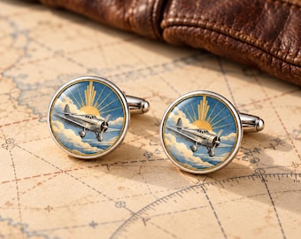 Könnte beinhalten: Ein Paar runde, silberfarbene Manschettenknöpfe mit einer detaillierten Abbildung eines alten Propellerflugzeugs, das durch einen blauen Himmel mit weißen Wolken und einem goldenen Sonnenstrahlenmotiv fliegt.