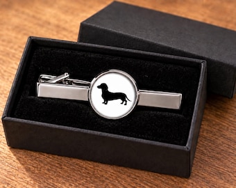Könnte beinhalten: Eine silberfarbene Krawattenklammer mit einem runden Mittelstück, das eine schwarze Silhouette eines Dackels auf weißem Hintergrund zeigt, präsentiert in einer schwarzen rechteckigen Geschenkbox mit schwarzem Samtfutter.