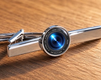 Könnte beinhalten: Eine silberfarbene Krawattenklammer aus Metall mit einem kreisförmigen Kameraobjektiv-Design auf der Vorderseite. Das Objektivelement zeigt blaues Glas mit dem Text ZOOM LENS, 24-70mm 1:2.8, USM, 82mm und LENS MADE IN JAPAN auf dem schwarzen Ring.