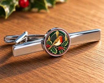 Könnte beinhalten: Eine silberfarbene Krawattenklammer mit einer runden Einlage. Die Einlage zeigt eine detaillierte Illustration eines Rotkehlchens auf einem Stechpalmenzweig mit roten Beeren vor einem dunkelgrünen Hintergrund mit goldenen Strahlen.