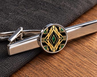 Könnte beinhalten: Eine silberfarbene Krawattenklammer aus Metall mit einem kreisförmigen Mittelstück, das ein geometrisches Muster in Schwarz, Gold und Grün aufweist. Das Design besteht aus symmetrischen Linien und Rautenformen in einem sich wiederholenden Motiv.