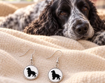 Könnte beinhalten: Ein Paar silberfarbene Ohrhänger mit runden Anhängern, die einen weißen Hintergrund und die schwarze Silhouette eines Spaniels zeigen. Die Ohrringe verfügen über klassische Hakenverschlüsse und einen polierten Metallrahmen um die Motive.