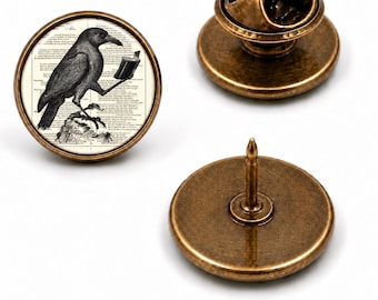 Könnte beinhalten: Eine runde Anstecknadel aus Bronze mit einer schwarz-weißen Illustration einer Krähe, die ein offenes Buch vor einem Hintergrund aus gedrucktem Text hält. Die Nadel hat eine Metallrückseite mit Stift und einem sicheren Verschluss.
