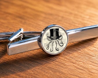 Könnte beinhalten: Eine silberfarbene Krawattenklammer mit einem runden Glaskabochon. Das Inlay zeigt eine schwarz-weiße Illustration eines Oktopus mit Zylinder vor einem Hintergrund aus antik wirkendem, bedrucktem Text auf cremefarbenem Papieruntergrund.
