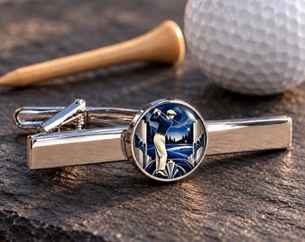 Könnte beinhalten: Eine silberfarbene Krawattenklammer mit einer runden Einlage, die einen Golfer in blauem Hemd und weißer Hose vor einer stilisierten Landschaft zeigt. Die Krawattenklammer liegt neben einem weißen Golfball und einem Tee aus Holz.