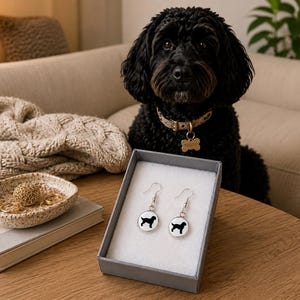 Op de afbeelding: Een paar zilverkleurige hangende oorbellen met een ronde witte hanger met een zwarte silhouet van een hond. De oorbellen worden gepresenteerd in een grijze geschenkdoos met een witte schuimrubberen inzet op een houten ondergrond.