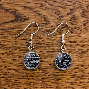 Puede incluir: Un par de pendientes de aro de plata con un dije redondo que presenta una ecuación matemática compleja impresa en negro.