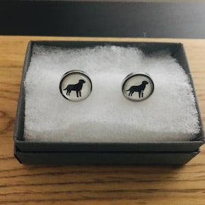 May include: A pair of silver stud earrings with a black silhouette of a dog on each earring. The earrings are in a white box with a gray lid.