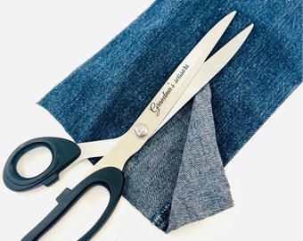 May include: Silver sewing scissors with black handles, resting on a piece of blue denim fabric. The scissors are engraved with the text "Grandma's scissors".