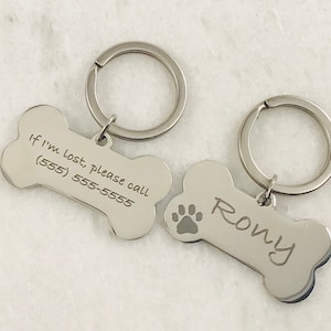 Puede incluir: Dos etiquetas para perros de hueso de plata con llaveros. Una etiqueta tiene el texto "Si me pierdo, por favor llama al (555) 555-5555" y la otra etiqueta tiene el nombre "Rony" grabado.