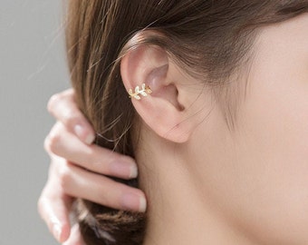 May include: A gold-colored ear cuff in the shape of a delicate leaf design. The jewelry piece is worn on the ear and is visible against the skin. The ear cuff is a stylish accessory.