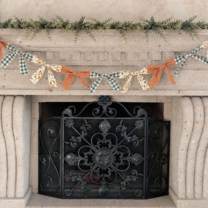 May include: A decorative garland with fabric bows in orange, green gingham, and floral patterns. The garland is draped across a stone fireplace mantel, with greenery above. The fireplace has a black metal screen.