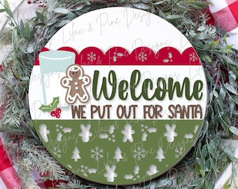 May include: A round wooden sign with a green, red, and white Christmas theme. The sign says "Welcome We Put Out For Santa" with a gingerbread man and a glass of milk.
