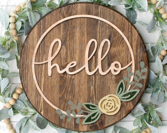 Könnte beinhalten: Ein rundes Holzschild mit dem Wort "hello" in einer kursiv geschriebenen Schriftart innerhalb eines Kreises. Das Schild ist mit einem floralen Design mit grünen Blättern und einer gelben Rose verziert.