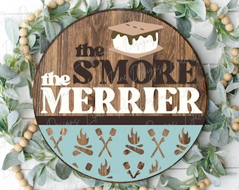 Könnte beinhalten: Ein rundes Holzschild mit dem Text "the s'more merrier" in weißen Buchstaben auf braunem Hintergrund. Das Schild ist mit einem blauen und weißen Muster aus Marshmallows, Feuer und Stöcken verziert.