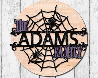 Puede incluir: Un letrero redondo de madera con un diseño de telaraña negra y el texto "The Adams Family" en letras moradas y negras. El letrero es perfecto para la decoración de Halloween.