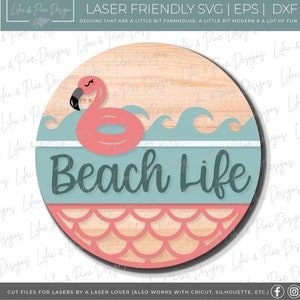 Puede incluir: Un letrero redondo de madera con un flamenco rosa flotando en una ola azul. El letrero dice "Beach Life" en una fuente verde azulada. La parte inferior del letrero tiene un patrón de escamas de sirena rosa.