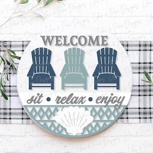 May include: A round wooden sign with a blue and white seashell design. The sign says "Welcome, sit relax enjoy" and features three blue and teal Adirondack chairs.