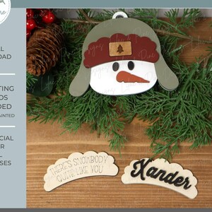 May include: A wooden snowman ornament with a green hat, brown ear flaps, and a carrot nose. The ornament has the text "There's Snowbody Quite Like You" and a name tag that says "Xander". The image also includes text that says "Laser & Cricut - SVG | EPS | DXF".