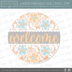 May include: A round wooden sign with a gray background and the word "welcome" in a yellow script font. The sign is decorated with a white cutout design of flowers and leaves. The background of the sign is a wood grain pattern.