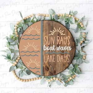 Op de afbeelding: Een rond houten bord met een gesplitst ontwerp. De linkerzijde is geschilderd in tinten bruin en blauw met een zon- en golfontwerp. De rechterzijde is van natuurlijk hout met de tekst "SUN RAYS boat waves LAKE DAYS".