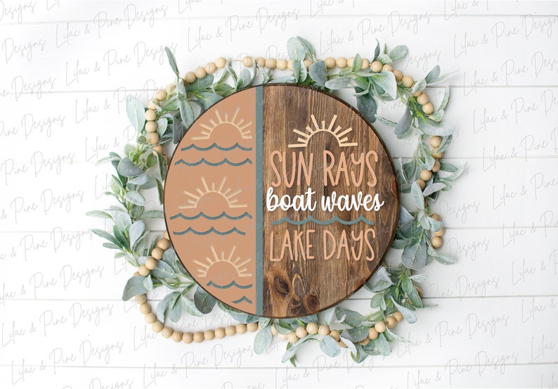 Op de afbeelding: Een rond houten bord met een gesplitst ontwerp. De linkerzijde is geschilderd in tinten bruin en blauw met een zon- en golfontwerp. De rechterzijde is van natuurlijk hout met de tekst "SUN RAYS boat waves LAKE DAYS".