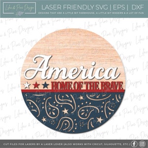 Puede incluir: Un letrero redondo de madera con un dise&ntilde;o patri&oacute;tico rojo, blanco y azul. El letrero dice "America Home of the Brave".