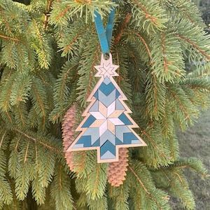 Op de afbeelding: Een houten kerstboomversiering met een geometrisch patroon in tinten teal en wit. De versiering hangt aan een blauw lint aan een groene dennentak.