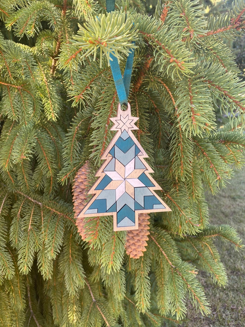 Op de afbeelding: Een houten kerstboomversiering met een geometrisch patroon in tinten teal en wit. De versiering hangt aan een blauw lint aan een groene dennentak.