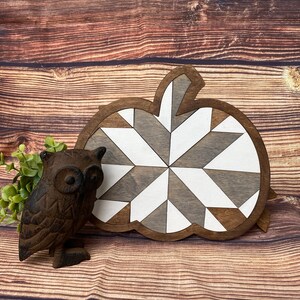 May include: A wooden pumpkin-shaped wall decor with a geometric pattern in shades of brown and white. The pumpkin is on a brown wooden background.