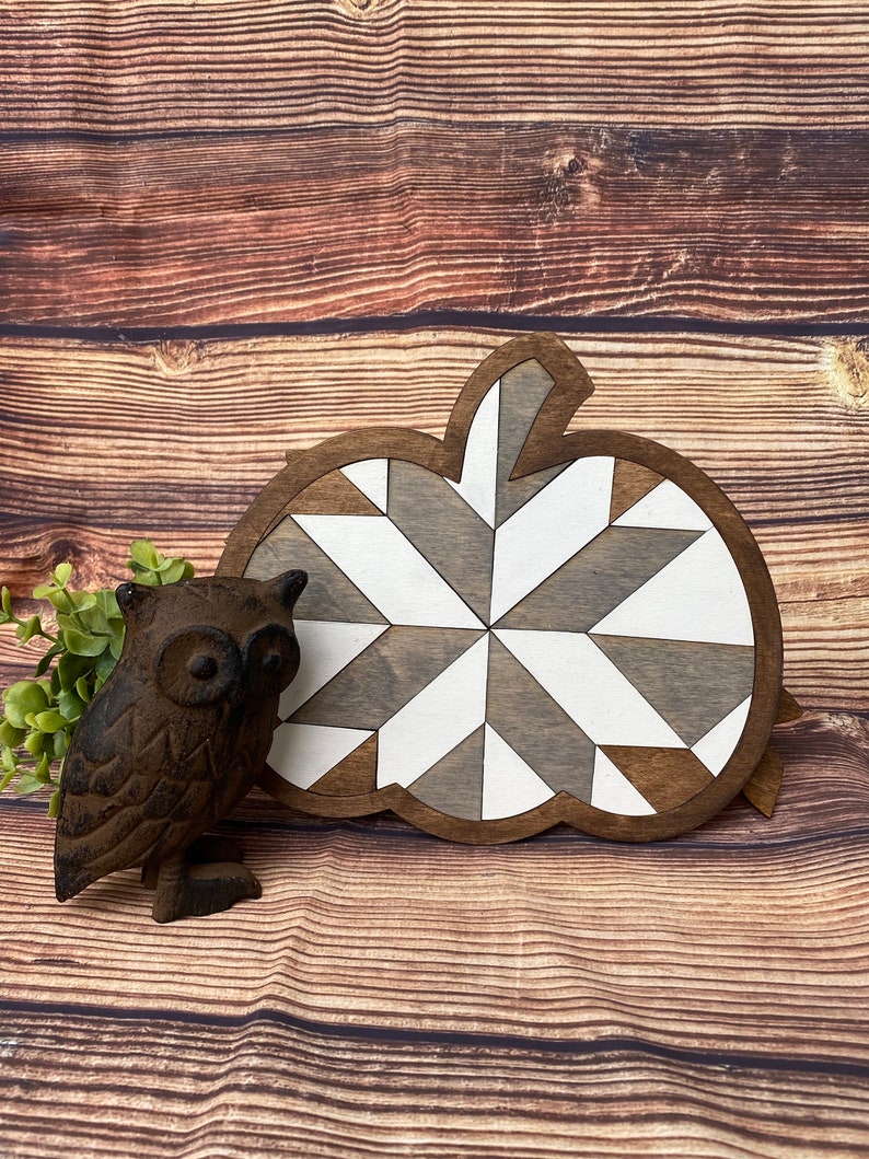 May include: A wooden pumpkin-shaped wall decor with a geometric pattern in shades of brown and white. The pumpkin is on a brown wooden background.
