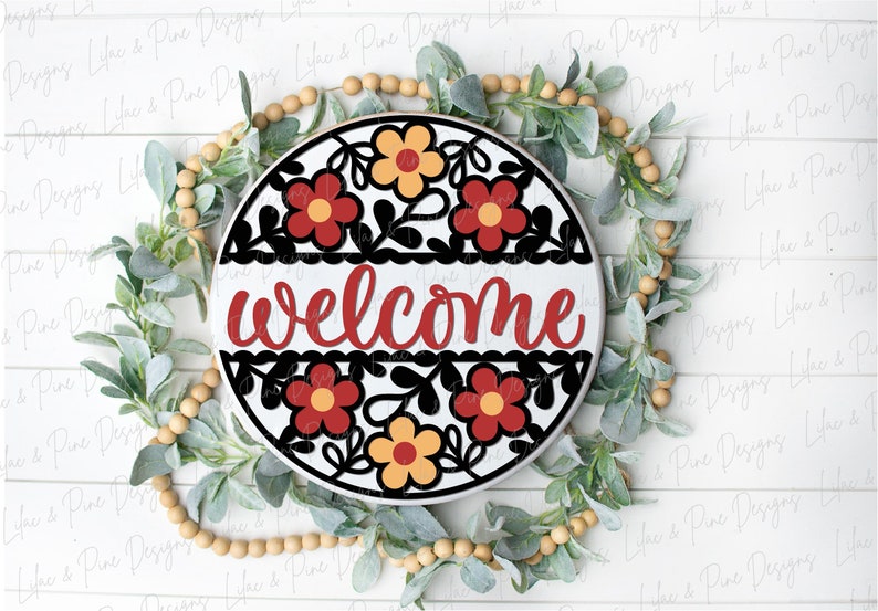 May include: A round wooden sign with a welcome message in red script. The sign is decorated with a black and white floral design with red and yellow flowers.