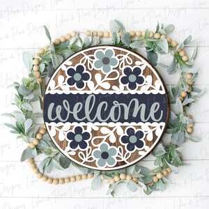 May include: A round wooden sign with a white background and a navy blue band across the middle. The sign says "welcome" in a gray script font. The sign is decorated with white and blue flowers and leaves.