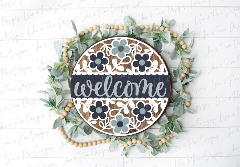May include: A round wooden sign with a white background and a navy blue band across the middle. The sign says "welcome" in a gray script font. The sign is decorated with white and blue flowers and leaves.