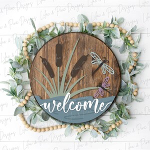 May include: A round wooden sign with a blue and green lake scene, featuring cattails and dragonflies. The sign says "welcome" in white cursive lettering.