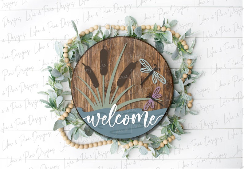 May include: A round wooden sign with a blue and green lake scene, featuring cattails and dragonflies. The sign says "welcome" in white cursive lettering.
