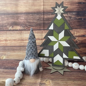 Op de afbeelding: Een zwart-witte gnome met een lange witte baard staat naast een groene, witte en grijze geometrische houten kerstboom. De boom heeft een ster bovenop en is omringd door een reeks witte kralen.