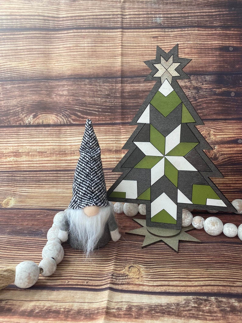 Op de afbeelding: Een zwart-witte gnome met een lange witte baard staat naast een groene, witte en grijze geometrische houten kerstboom. De boom heeft een ster bovenop en is omringd door een reeks witte kralen.