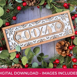 May include: A rectangular wooden sign with the word "COZY" in white, surrounded by a white snowflake and geometric design. The sign is set against a backdrop of greenery, red berries, and pinecones, creating a festive, seasonal theme.