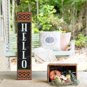 May include: A wooden sign with the word "HELLO" in white letters on a black background. The sign has a brown and orange patterned border with pumpkins and leaves. The sign is leaning against a white porch swing with a pillow that has a gray pumpkin on it.