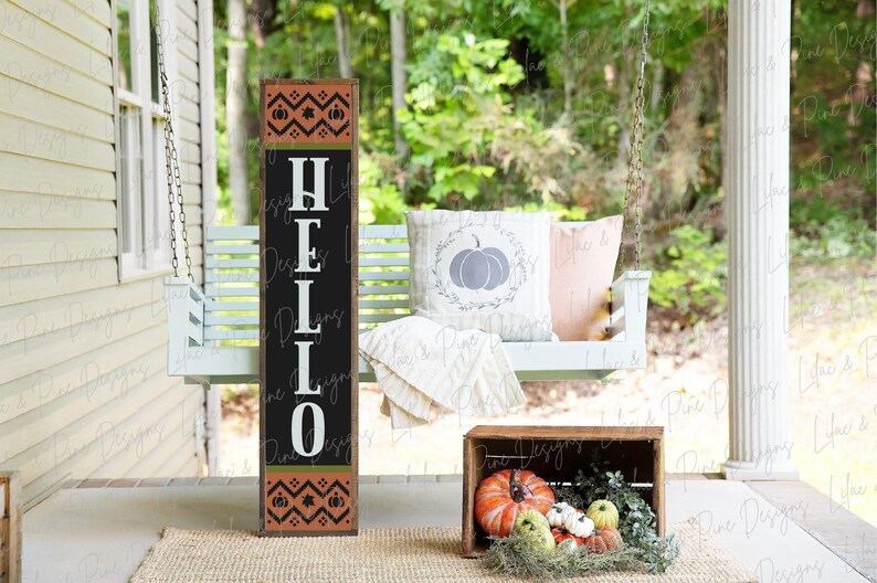 May include: A wooden sign with the word "HELLO" in white letters on a black background. The sign has a brown and orange patterned border with pumpkins and leaves. The sign is leaning against a white porch swing with a pillow that has a gray pumpkin on it.